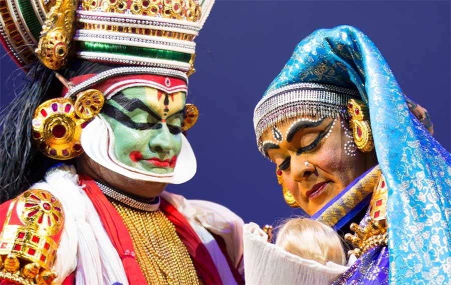 Two individuals with face paint and elaborate headdresses are looking down at a doll during a performance. They are wearing brightly coloured costumes