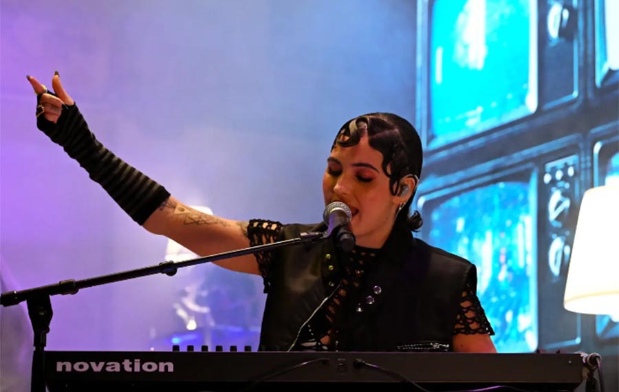 A young girl with dark hair is singing into a microphone, looking down and playing a keyboard.