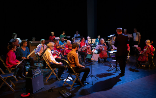An orchestra made up of around 30 people are sat down in a semi circle with music stands in front of them. They are all reading music. The conductor is stood at the front, facing the orchestra