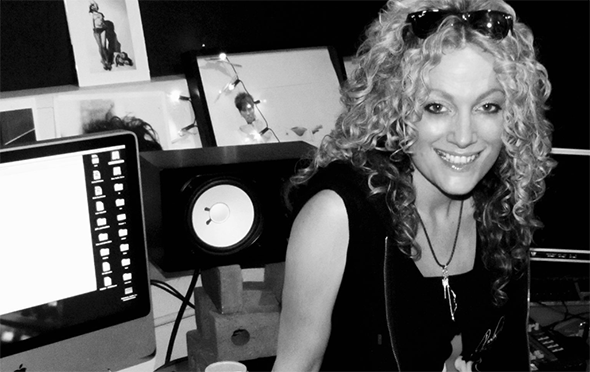 A young woman with blonde, curly hair is smiling directly into the camera. She is wearing a necklace. Behind her is an Apple Mac with a programme open