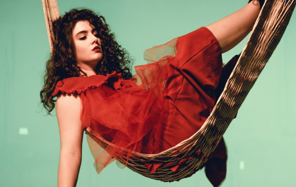 A young woman is seen lying down on a hammock, looking past the camera. She is wearing a burnt orange coloured dress and has long, brown, curly hair