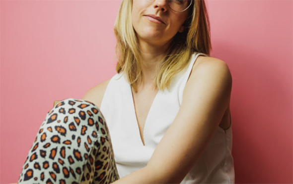 Shows a young woman with straight blonde hair sat down with one knee up. Her hand is across her knee and she is smiling slightly. She is wearing a white vest and leopard print trousers