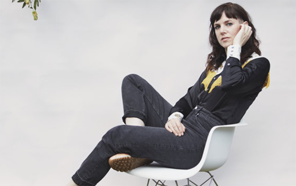 A young woman is sat down on a white chair, with one leg crossed under the other. She has her hand up to her ear and she is wearing black jeans and a black shirt. She has middle-length brown hair
