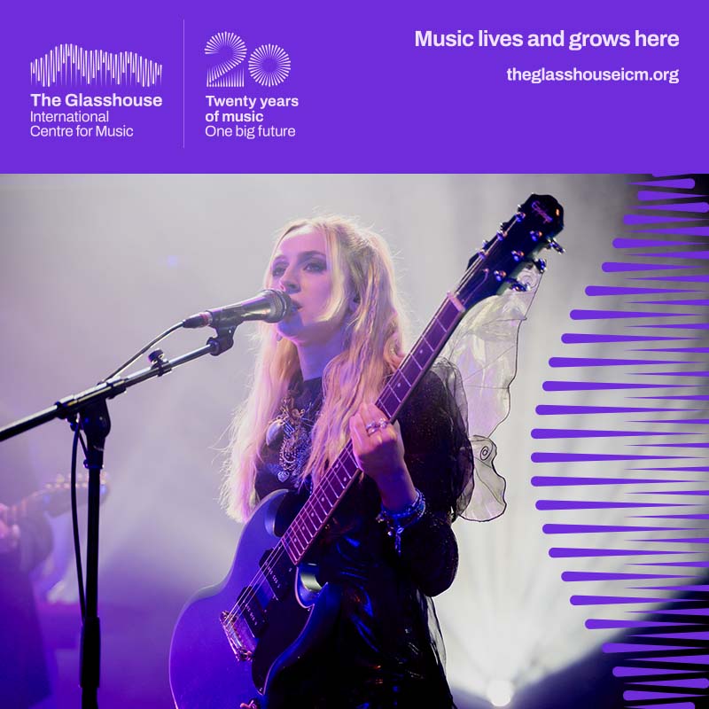 Lizzie Esau: A woman with blonde hair wearing a black dress and fairy wings. She's playing a guitar and singing into a microphone.