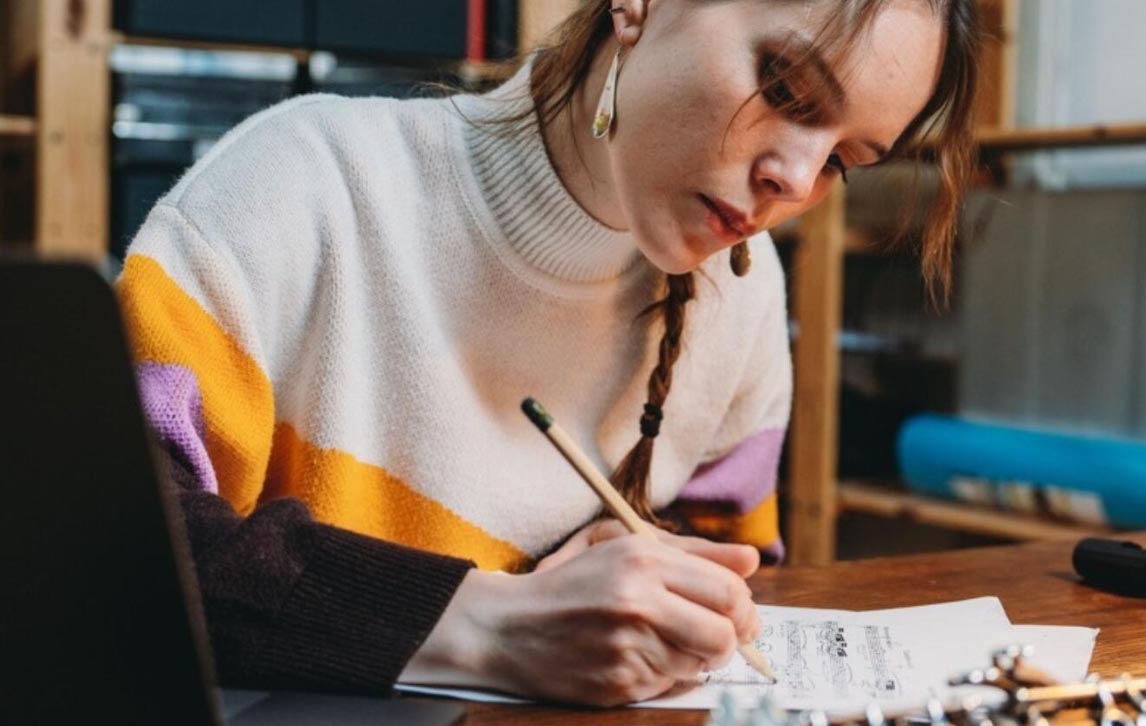 A young girl in a beige, pink and orange jumper. She is writing notation on manuscript paper.