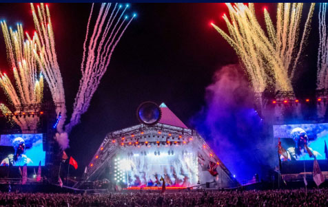 A festival stage with two screens either side showing the performance. A large crowd is watching the stage and multicoloured fireworks are going off behind each screen.