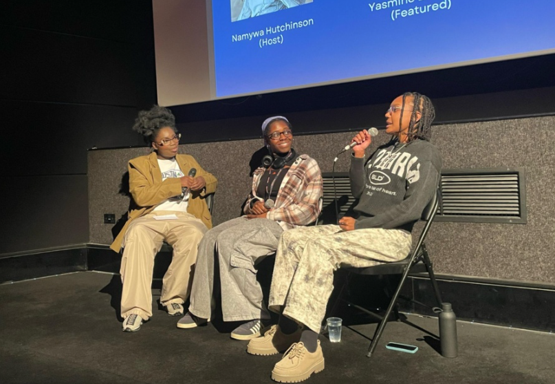 Three young women are sat down in a line. They are all holding microphones. The young woman to the right is speaking into her microphone whilst giving a presentation. They are all smiling