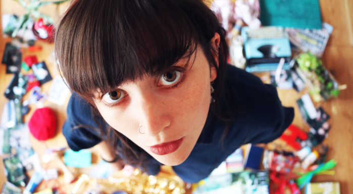 A young woman who is stood up with brown hair, a fringe and brown eyes stares straight into the camera. She is wearing a blue top. The background is blurry but showcases a messy floor with lots of arts and crafts surrounding the girl.
