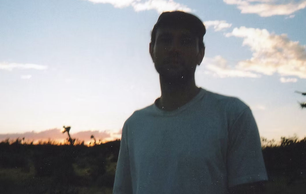 A young man in a white, short sleeved T shirt is standing looking directly into the camera. There is a sunset behind him and the image is slightly blurred