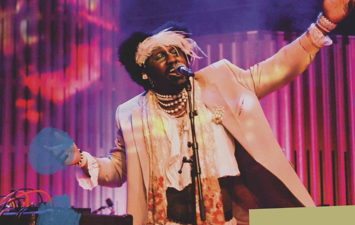 A young man is seen standing and singing into a microphone. He has both arms in the air and is wearing a dusky pink suit with a pearl necklace and satin scarf. He is also wearing a feather headband