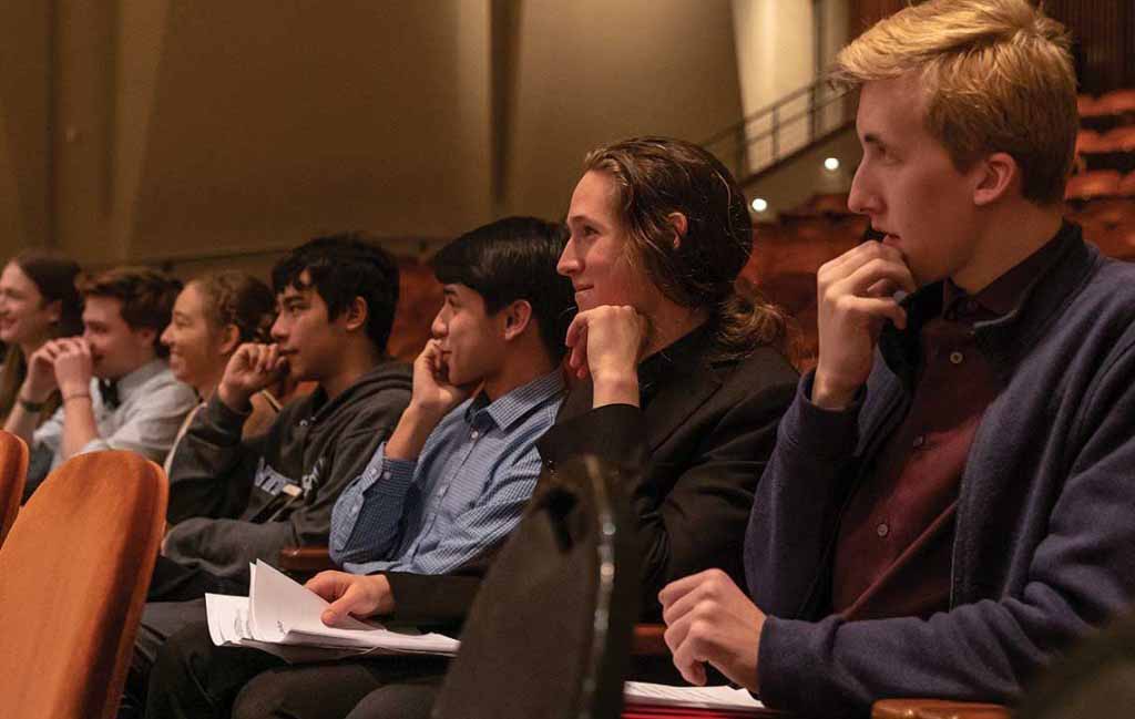 Seven young people - five male and two female are sat in a music auditorium. They are looking in the same direction as if it were a lecture. The five males all have their right hand to their face and all the individuals are smiling