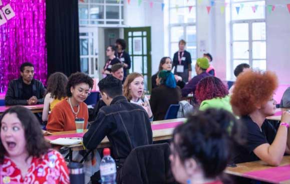 Approximately seventeen young people are shown. They are in a classroom setting and are talking to each other. Some are closer to the camera and others are in the background. This is an action shot