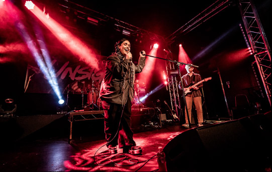 A young girl with a brown jacket is singing into a microphone on a stage. A young boy is stood behind her playing electric guitar. There is red lighting on the stage