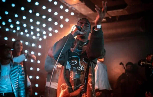 A young man is seen waving to the camera and performing into a microphone. There are spotlights behind him