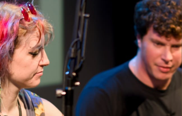 A young woman is seen to the left of the image looking down. She is wearing pink sunglasses and has pink hair. A young man is to the right of the image looking down too. He has brown curly hair and wearing a black t shirt