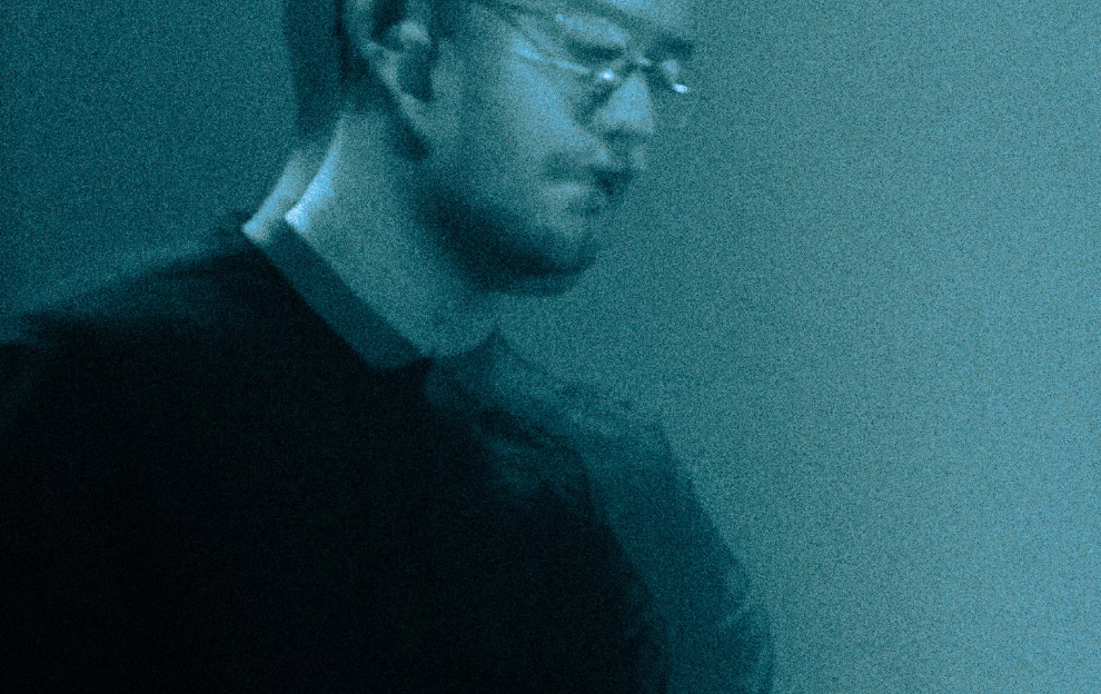 A young man is wearing glasses and looking down. He is wearing a black collared shirt and there is blue hazed lighting behind him