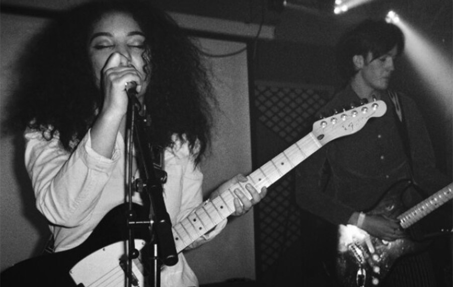 A young woman is playing an electric guitar and singing into a microphone. She has long, dark hair and wearing a white shirt. There is a young man stood behind her also playing electric guitar
