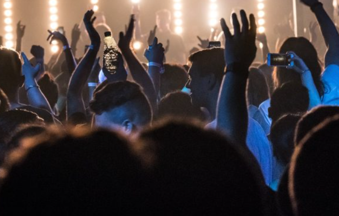 A crowd of people are seen at a concert with their hands in the air looking forward at the band on stage