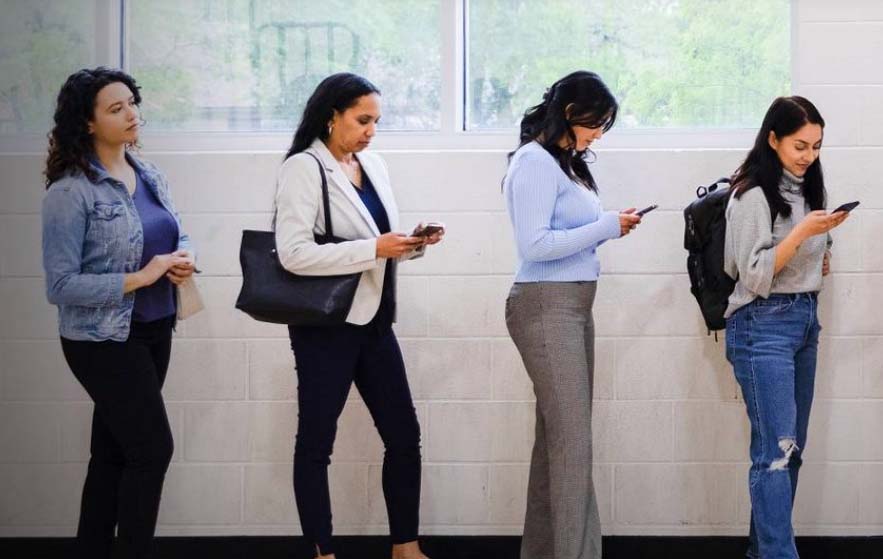 Four women are seen stood in a line on their phones. They are all wearing jeans and blazers