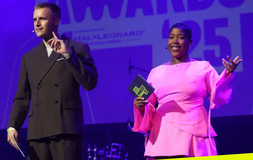 Two hosts are seen with head microphones on, presenting on a stage. One lady is wearing a bright pink dress and the other man is wearing a black suit