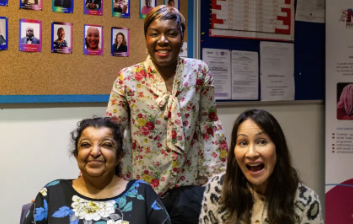 Three women are smiling directly into the camera. Two are sat down and one is stood behind them. They are all wearing bright coloured patterned shirts