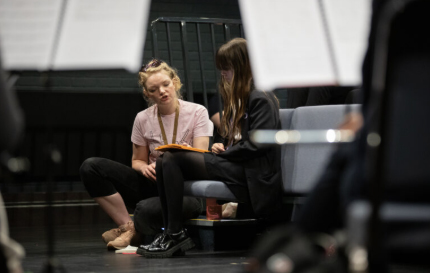 Two young women are sat down looking at a notepad. One is wearing a pink t shirt and the other is wearing a black jacket