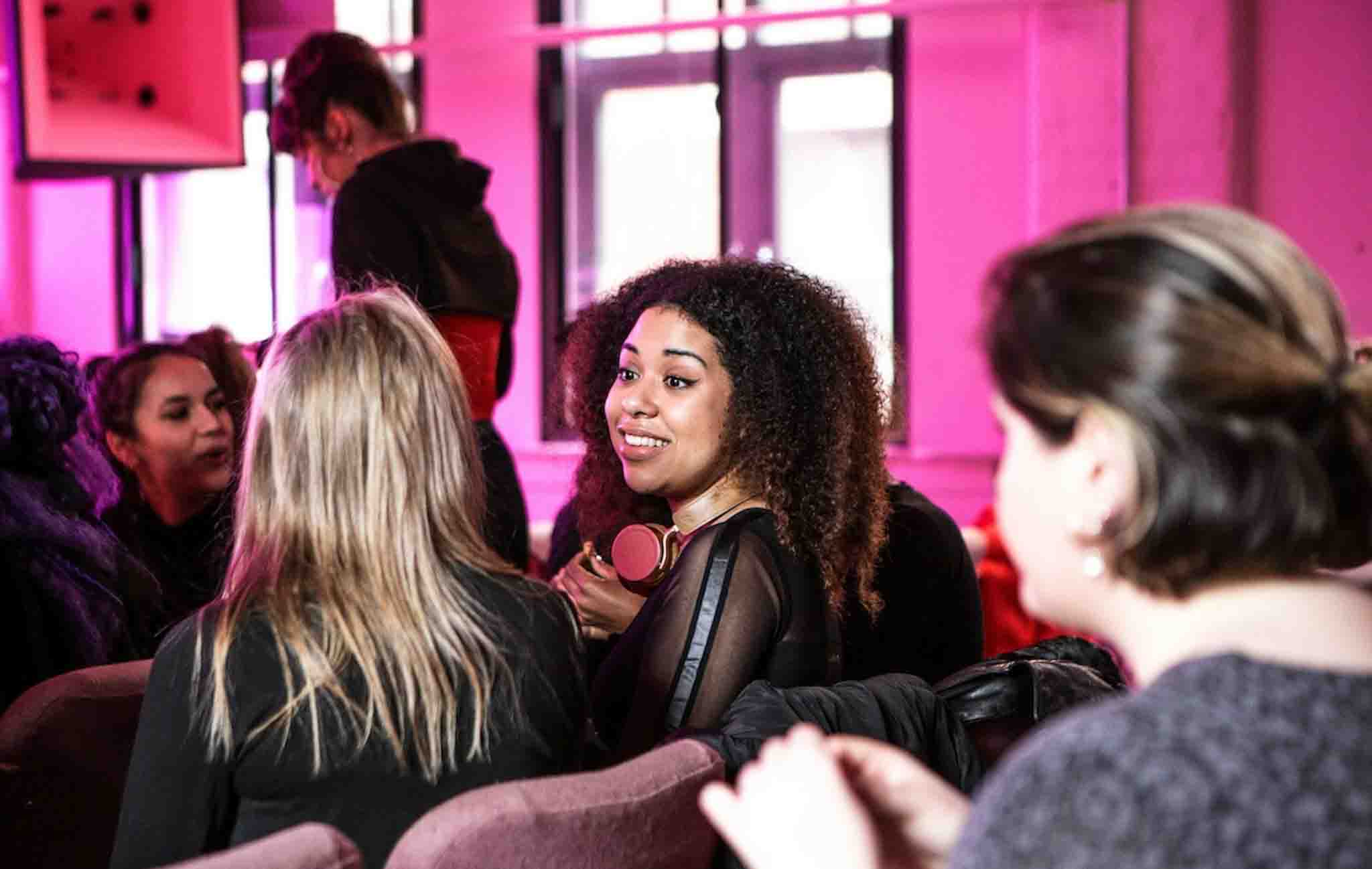 Three people are seen in the image. The young woman in the centre is holding a microphone and interviewing another young woman to her left