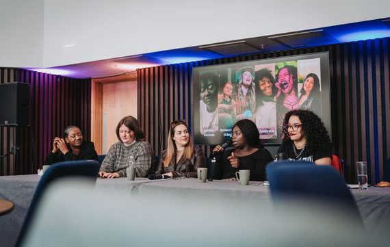 Five women are sat on a panel. There is a screen behind them with images of various women projected