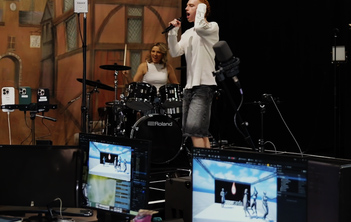 An individual is seen on a stage singing into a microphone. Another young woman is sat behind them playing the drums