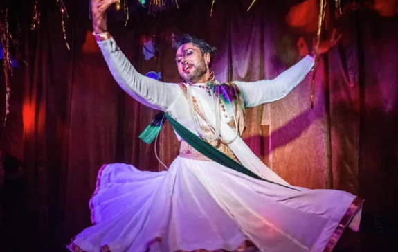 A performer is on stage with their hands in the air. They are wearing a white long dress and there is multicoloured lighting behind