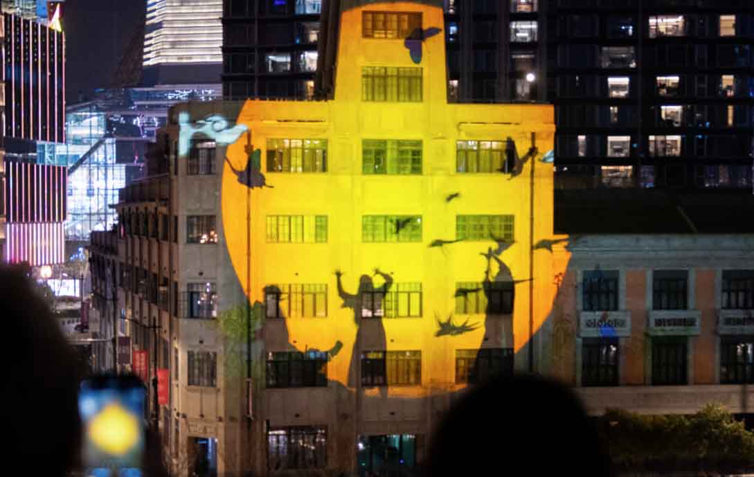 Shadow figures are projected onto a building, with yellow lighting
