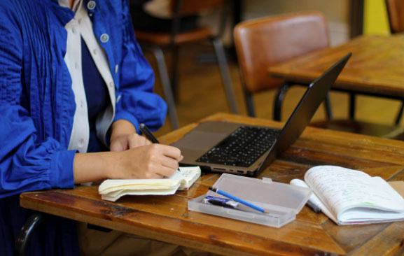 Someone is writing into a notepad and wearing a blue shirt. There is also a laptop next to them