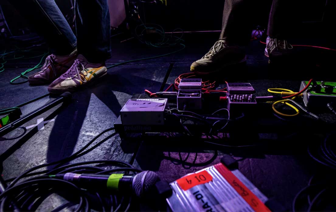 A pedal board is on the floor with wires coming out. There is also a microphone laid next to it on the floor