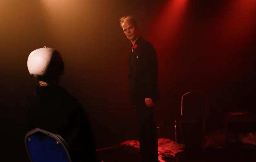 A man stands in the centre of a stage looking into the camera. He is backlit by red lighting