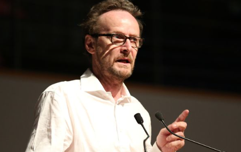 A man is talking into a microphone. He is wearing a white shirt and glasses