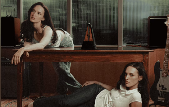 One young woman is laid across a table staring off to the left, and another is laid below her on the floor looking directly into the camera