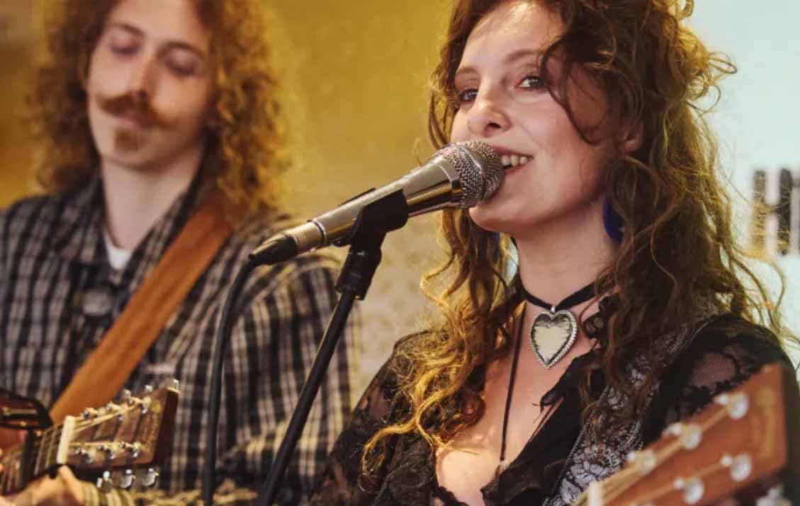A young woman with brown curly hair is singing into a microphone and playing an acoustic guitar. A young guy is stood behind her, just out of focus
