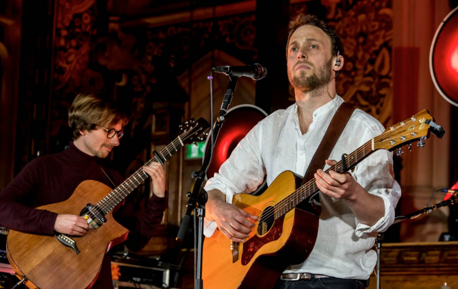 Two male performers are on stage both playing acoustic guitars