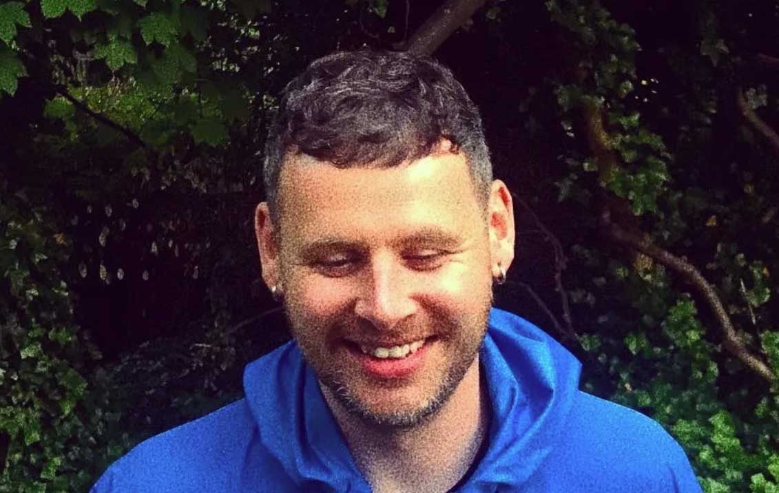 A man with short, dark coloured hair is staring down at the floor and smiling. He is wearing a blue hoodie