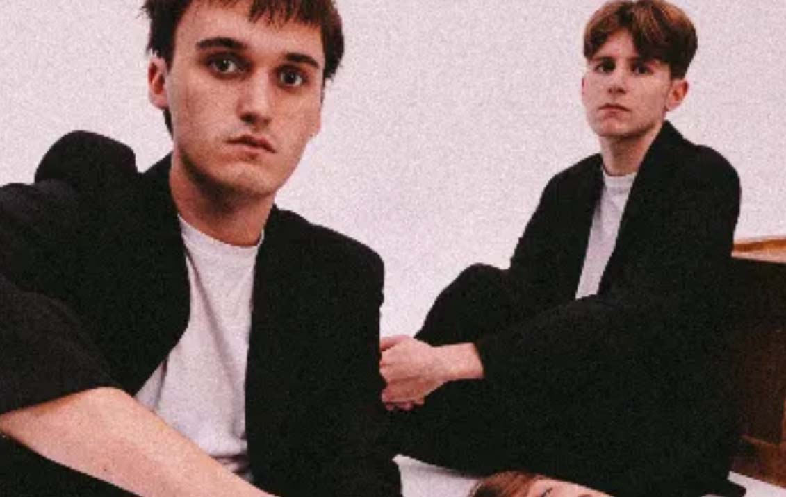 Two young men are sat down in black and white outfits staring straight into the camera. They both have brown hair