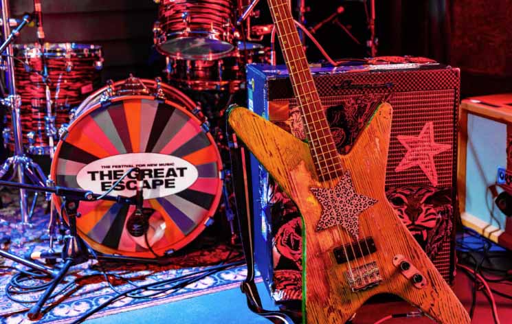 An electric guitar is propped up against an amp and a bass drum is in the background