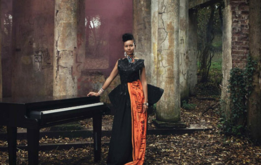 A young woman is stood with her hand on a grand piano. She is wearing a long black and orange dress