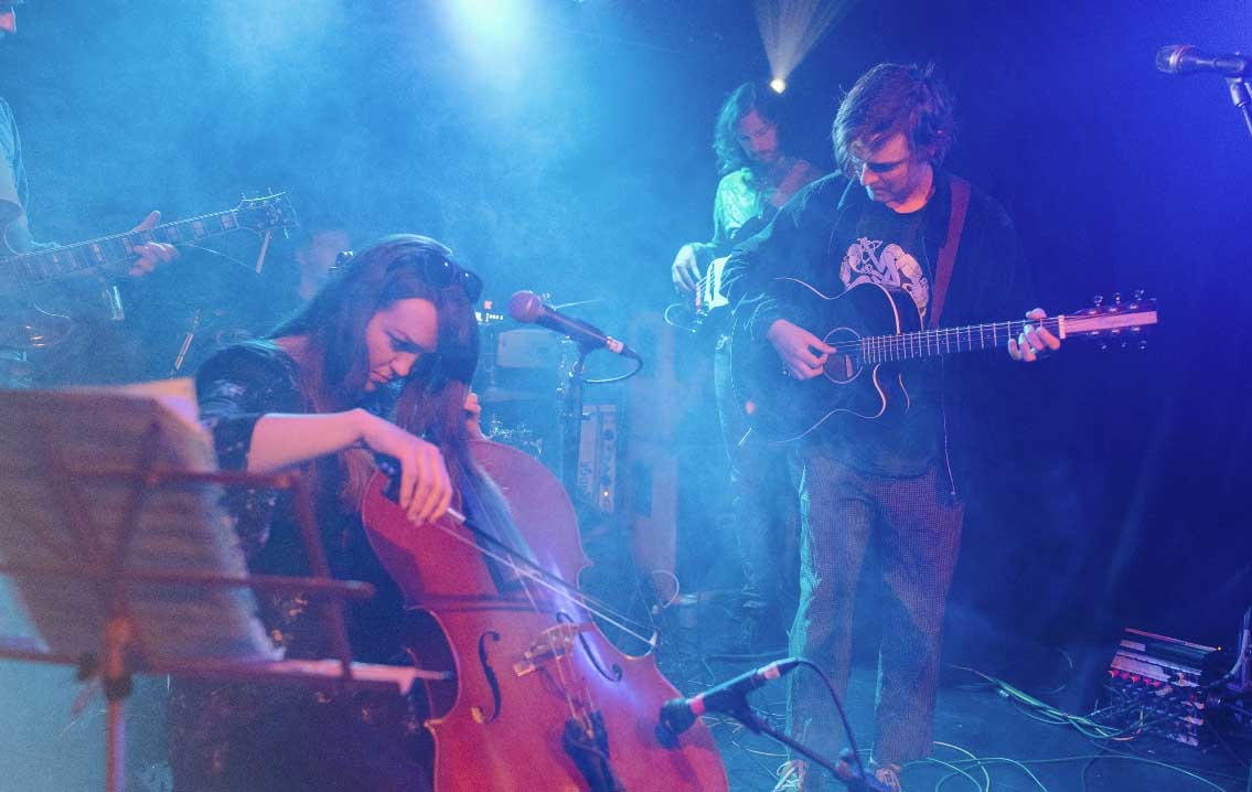 Three people are playing instruments on stage: one playing a double bass, one playing a bass guitar, and the final person one playing acoustic guitar