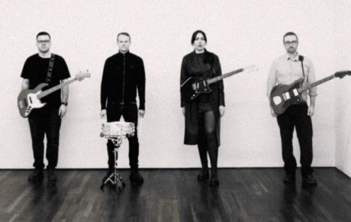 Four people are stood in a line with different instruments on them - three electric guitars and a snare drum