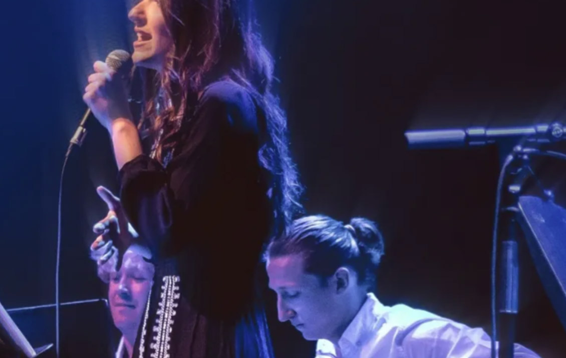 Three people are performing on stage: a young woman with long brown hair in the centre singing into a microphone, two young men sat beside her
