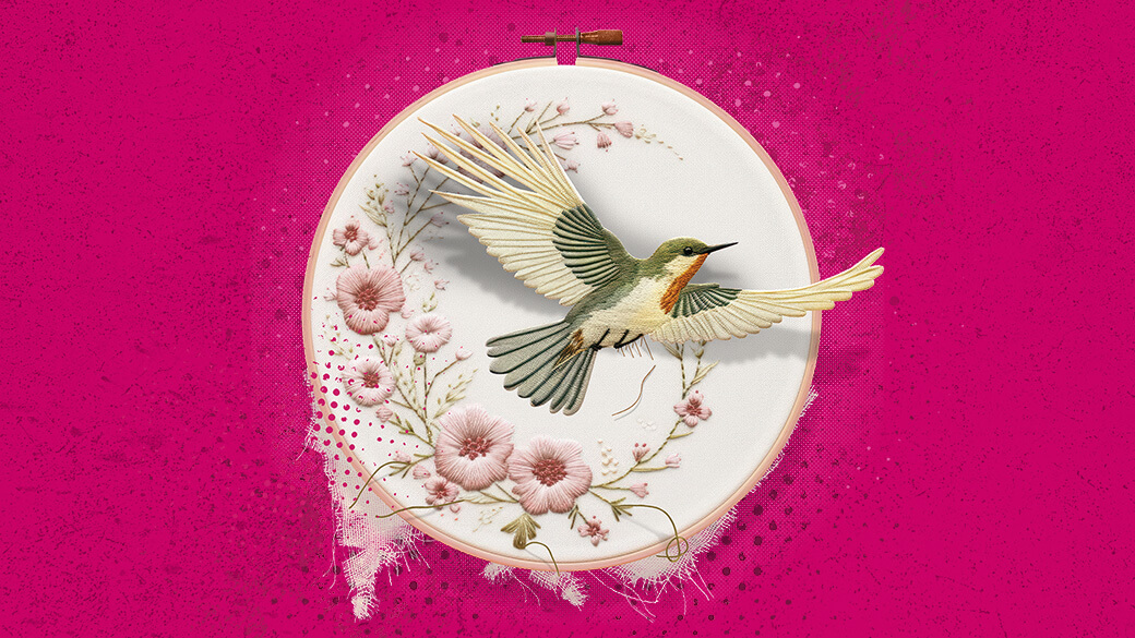 A white and gray bird hovering over an embroidery hoop with cross-stitched pink flowers on a bright pink background 