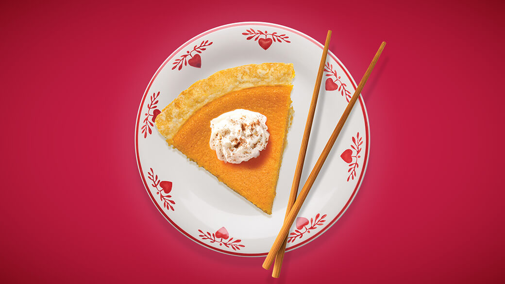 A piece of pumpkin pie is placed on a white plate with hearts around the edges. Their are chopsticks crossed on the side of the plate. The image is set against a red background.