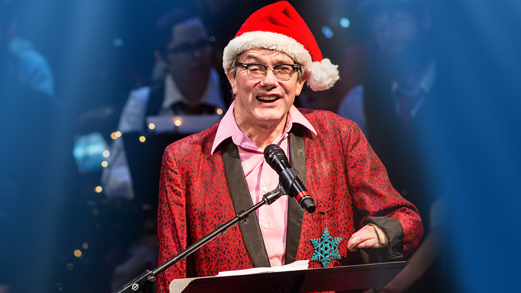 Playwright and humorist Kevin Kling performs onstage wearing a Santa hat, pink shirt and red blazer with a teal snowflake. Musicians and twinkle lights are visible in the background.