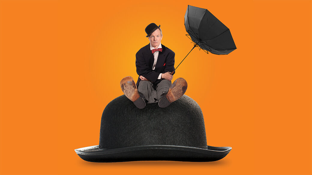 Actor Bill Irwin sits atop an oversized black bowler hat. He looks directly at the camera with a serious expression while holding an inverted black umbrella and wearing an askew bowler hat, formal suit and oversized clown shoes.