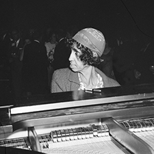 A close up black and white photograph of "Sweet" Emma Barrett playing the piano during a performance at an unidentified venue. She wears an embroidered skull cap that reads "Emma Bell Gal".